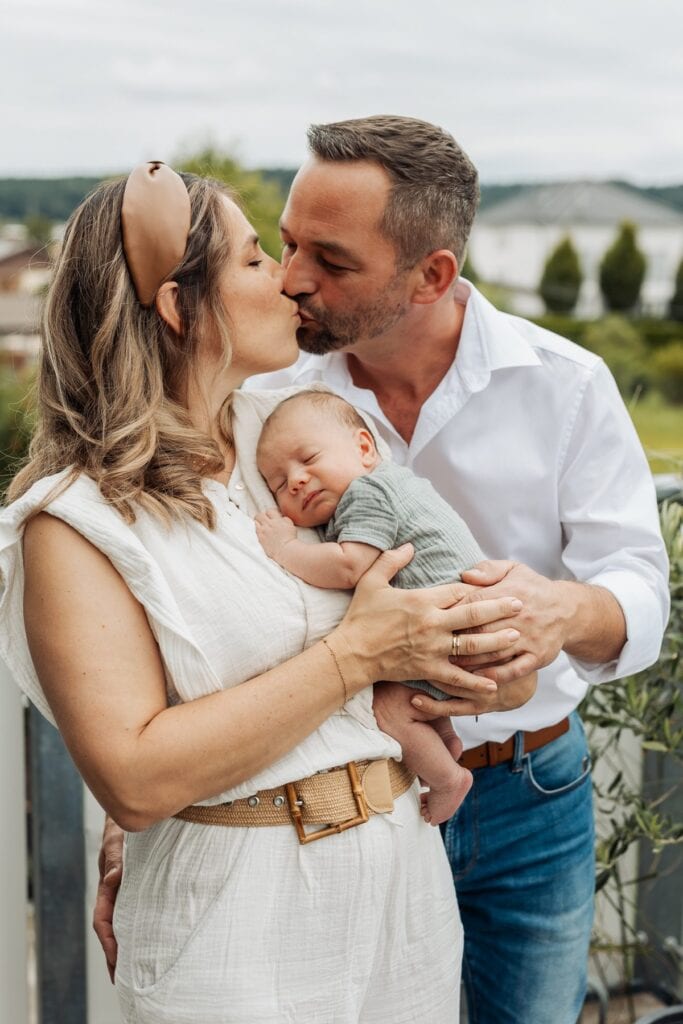 Ein Mann und eine Frau küssen ihr Baby, während sie auf einem Balkon stehen.