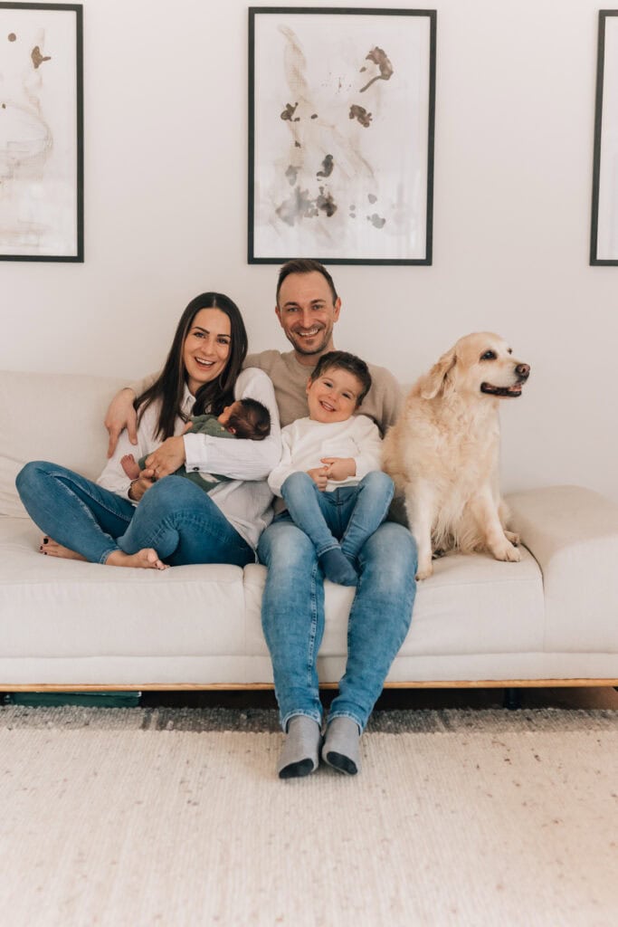 Eine vierköpfige Familie, darunter ein Baby und ein Kleinkind, sitzt mit einem Golden Retriever auf einem Sofa und lächelt in die Kamera. An der Wand hinter ihnen hängen zwei gerahmte Kunstwerke.