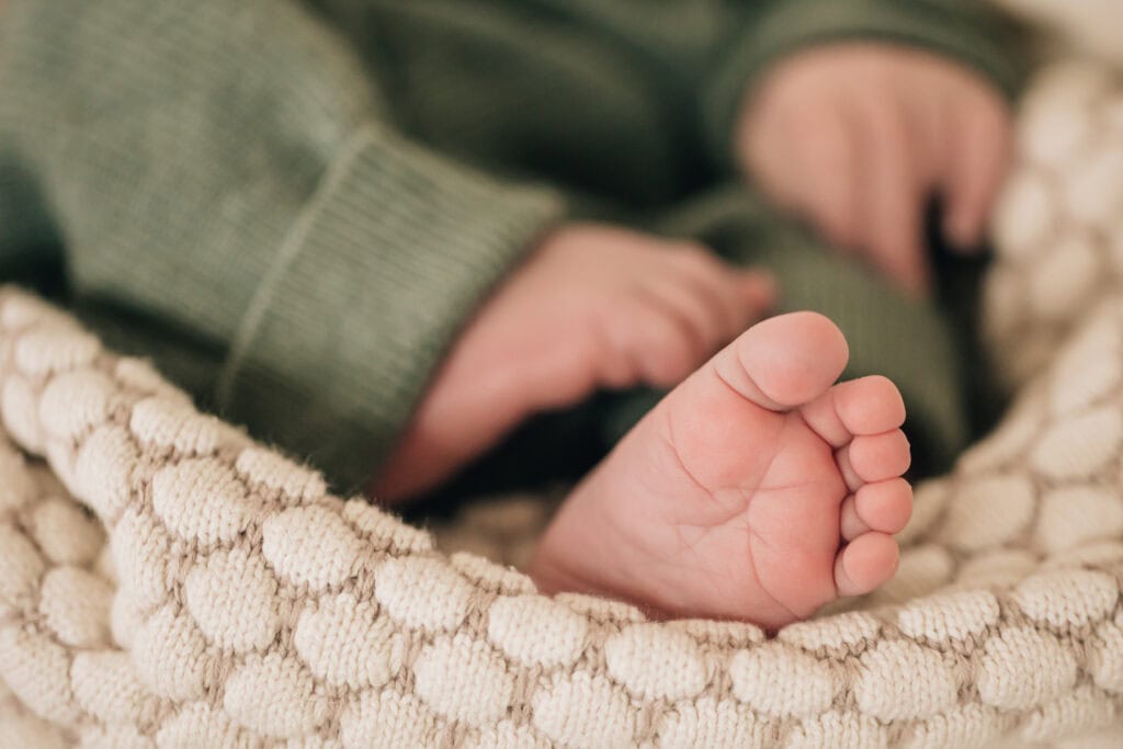 Nahaufnahme eines Babyfußes, der aus einem grünen Strampler hervorschaut, der auf einer strukturierten, cremefarbenen Decke ruht.