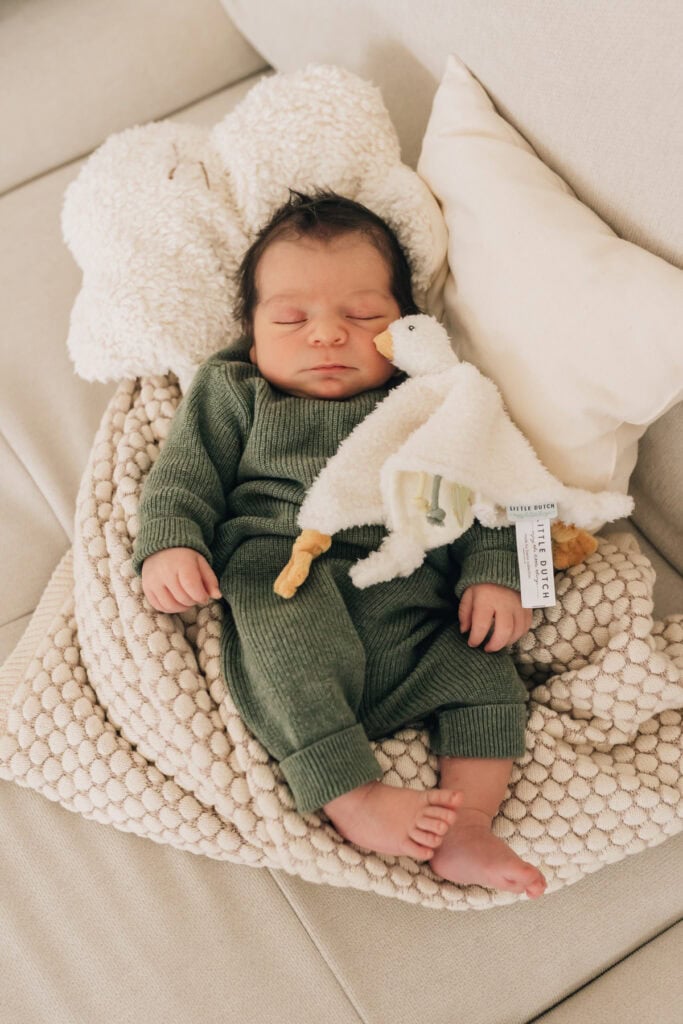 Ein Baby schläft auf einer strukturierten Decke, hat eine Stoffente unter dem Arm und ist in ein grünes Strickoutfit gehüllt und lehnt an einem Kissen.