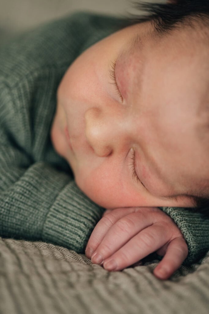 Nahaufnahme eines schlafenden Babys mit geschlossenen Augen, das auf dem Arm ruht und einen grünen Pullover trägt.