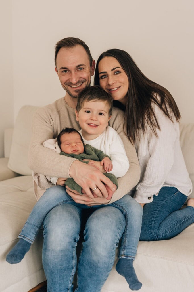 Eine vierköpfige Familie sitzt auf einem Sofa. Vater und Mutter sitzen lächelnd auf beiden Seiten, während der Vater ihr kleines Kind und ein Neugeborenes im Arm hält.