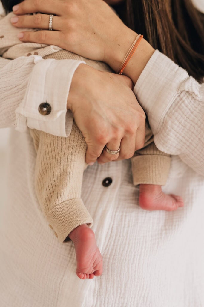 Eine Person in einem weißen Langarmshirt hält ein kleines Baby in beigefarbener Kleidung. Die winzigen Füße des Babys sind sichtbar. Die Person trägt einen Ring und ein orangefarbenes Armband.