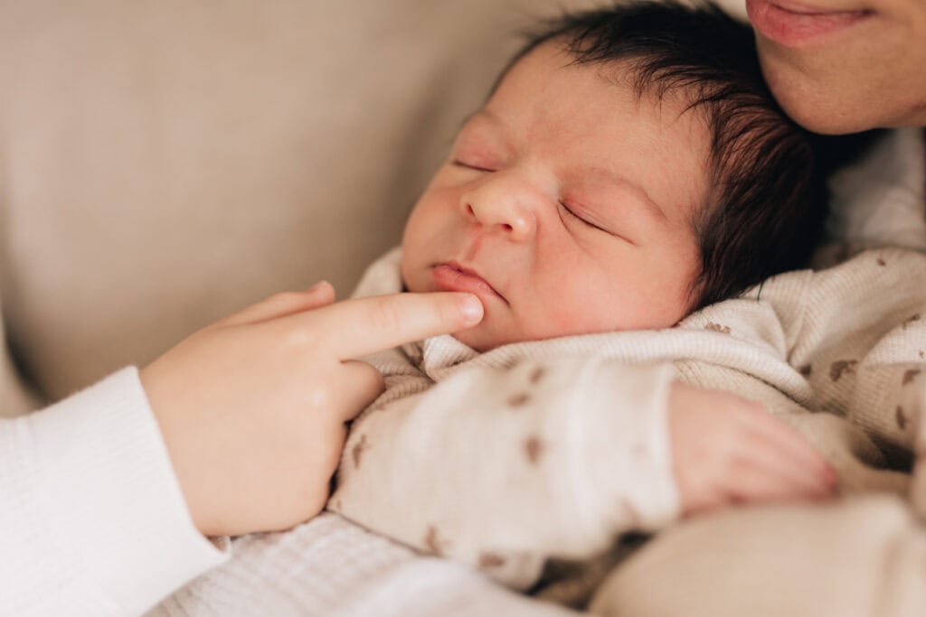 Eine Nahaufnahme eines Babys, das in den Armen einer Person schläft, während eine andere Person sanft das Kinn des Babys berührt.