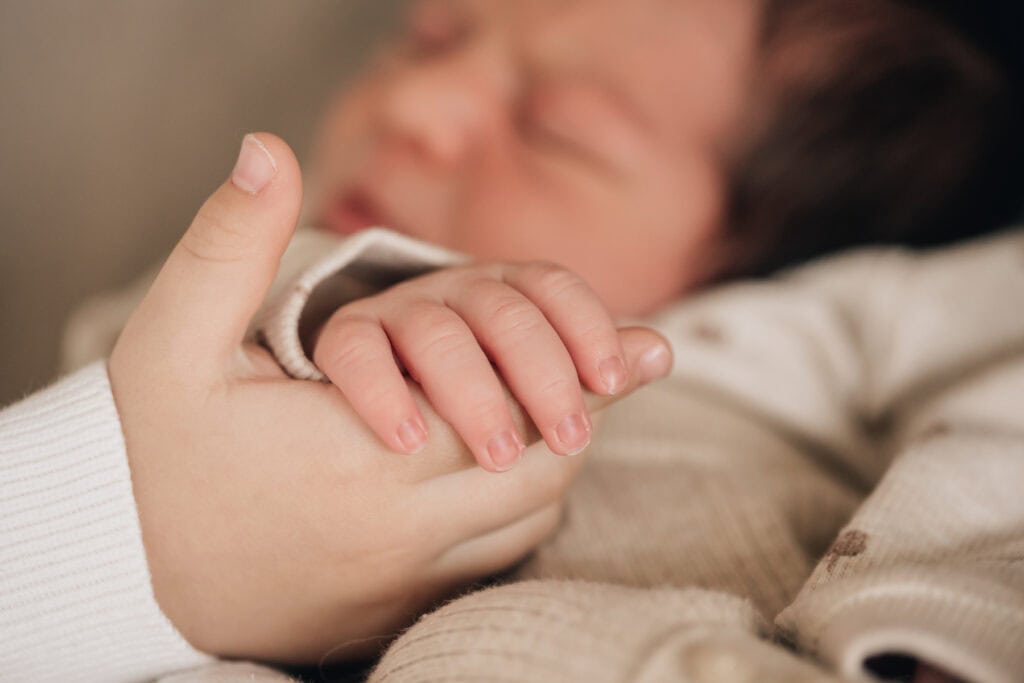 Nahaufnahme der Hand eines älteren Kindes, das sanft die Hand eines Neugeborenen hält. Beide tragen helle Kleidung.