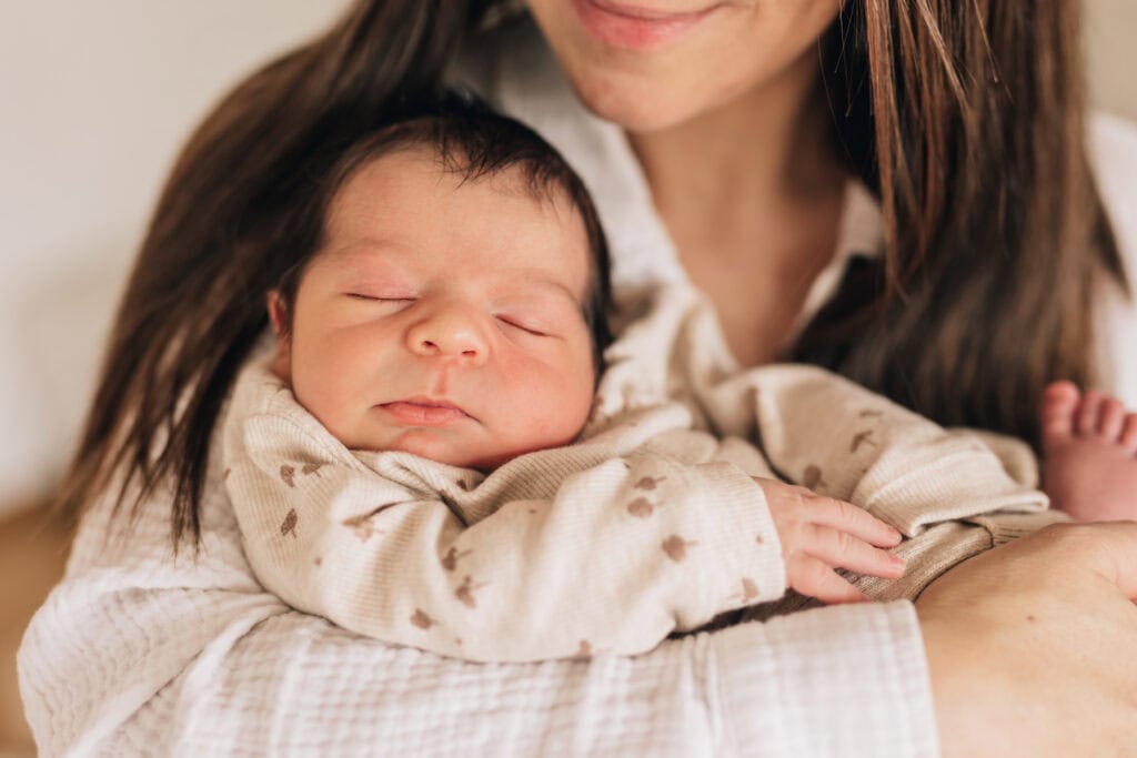 Eine Frau hält ein schlafendes Baby an ihrer Brust. Das Baby trägt ein helles Outfit mit kleinen Mustern.