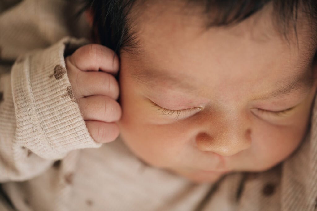 Nahaufnahme eines schlafenden Babys mit einer Hand in der Nähe des Gesichts, das ein beiges Outfit mit kleinen dunklen Flecken trägt.