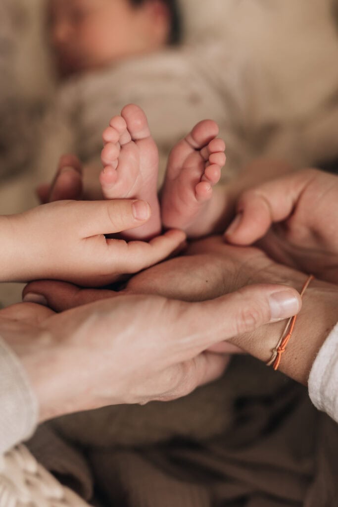 Nahaufnahme von mehreren Händen, die sanft die Füße eines Babys halten, wobei der Körper des Babys im Hintergrund unscharf ist.