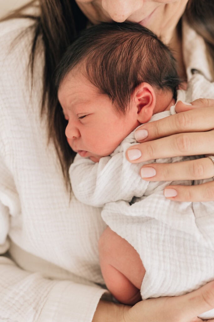 Ein Neugeborenes liegt an der Brust eines Erwachsenen, eingewickelt in eine weiße Decke, und die Hand des Erwachsenen hält sanft den Rücken des Babys.