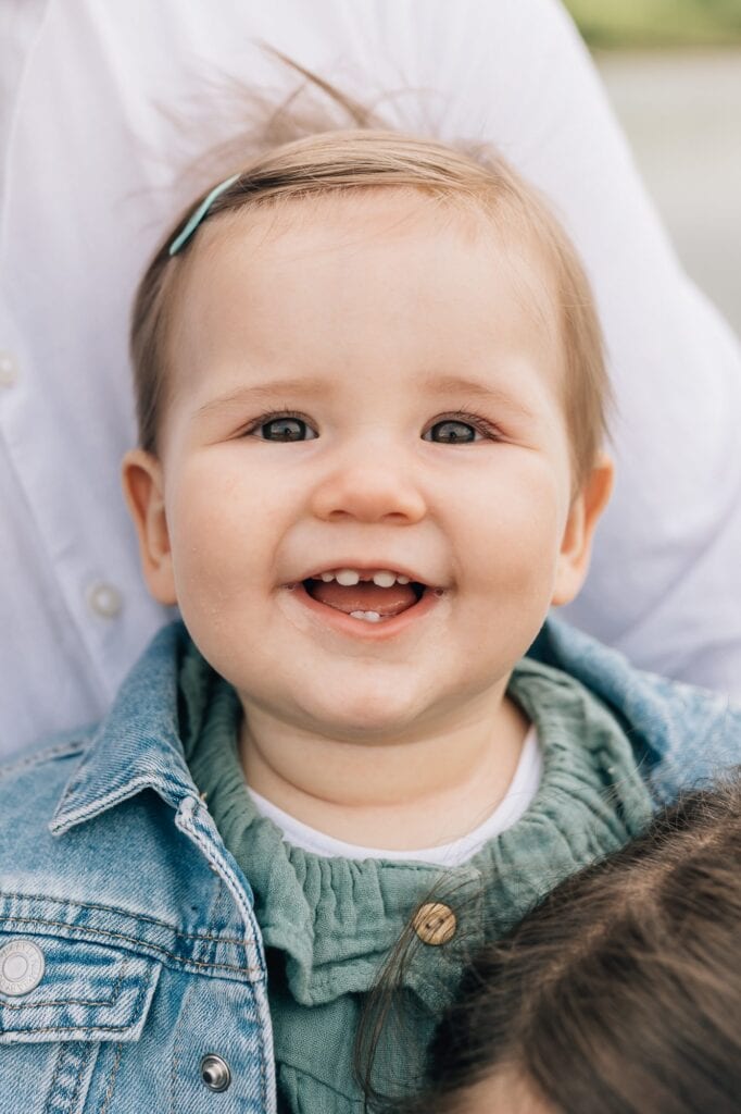 Ein Baby mit kurzen Haaren und einer Haarspange lächelt. Es trägt eine Jeansjacke über einem grünen Pullover.