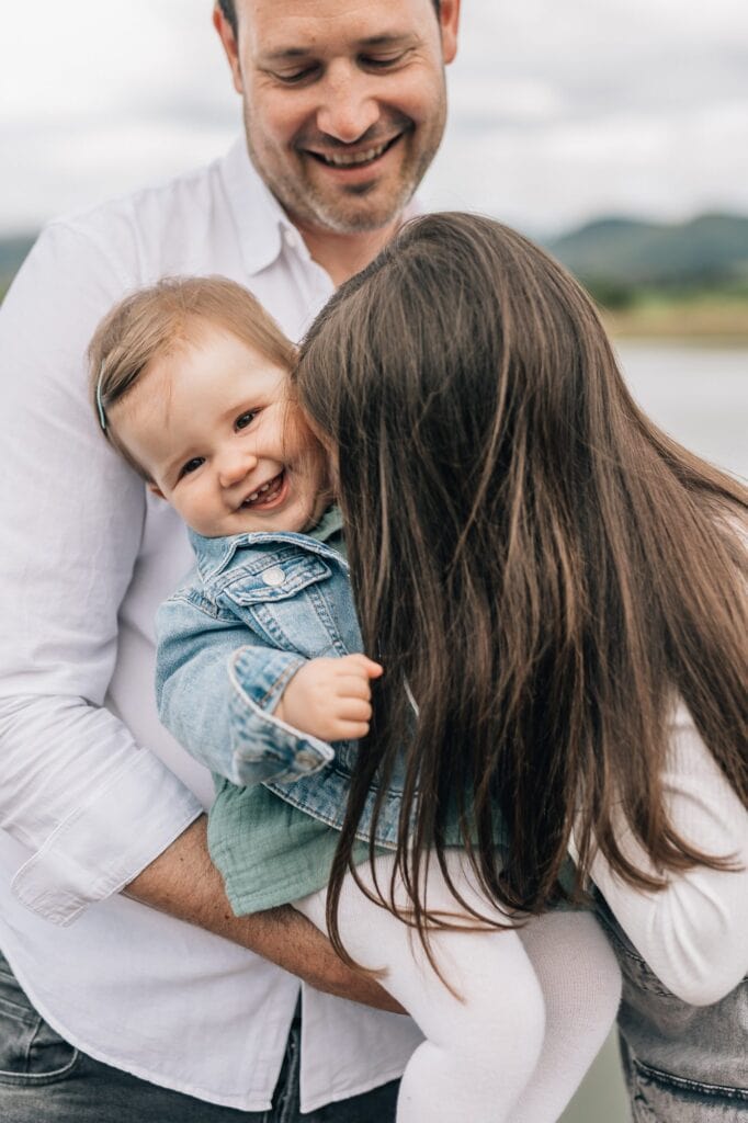 Ein Mann lächelt, während er ein Baby und ein Kind mit langen dunklen Haaren hält. Das Baby, das eine Jeansjacke trägt, lächelt freudig und wird teilweise von dem Kind vor ihm verdeckt.
