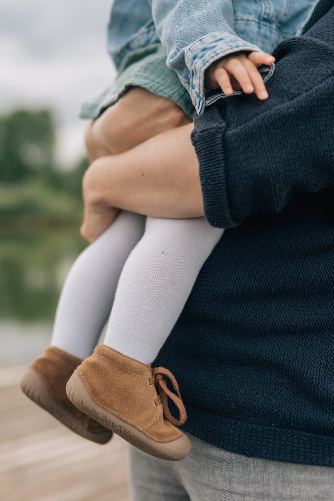 Ein Erwachsener hält ein kleines Kind, das weiße Strumpfhosen, braune Schuhe, ein grünes Kleid und eine Jeansjacke trägt. Von dem dunkelblauen Pullover des Erwachsenen ist nur der untere Teil zu sehen.