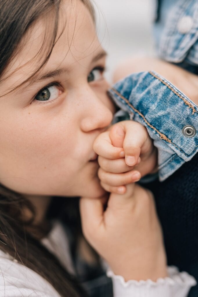 Ein Kind mit braunen Haaren und grünen Augen hält die Hand eines Babys vor den Mund. Das Baby trägt ein blaues Jeanskleidungsstück.