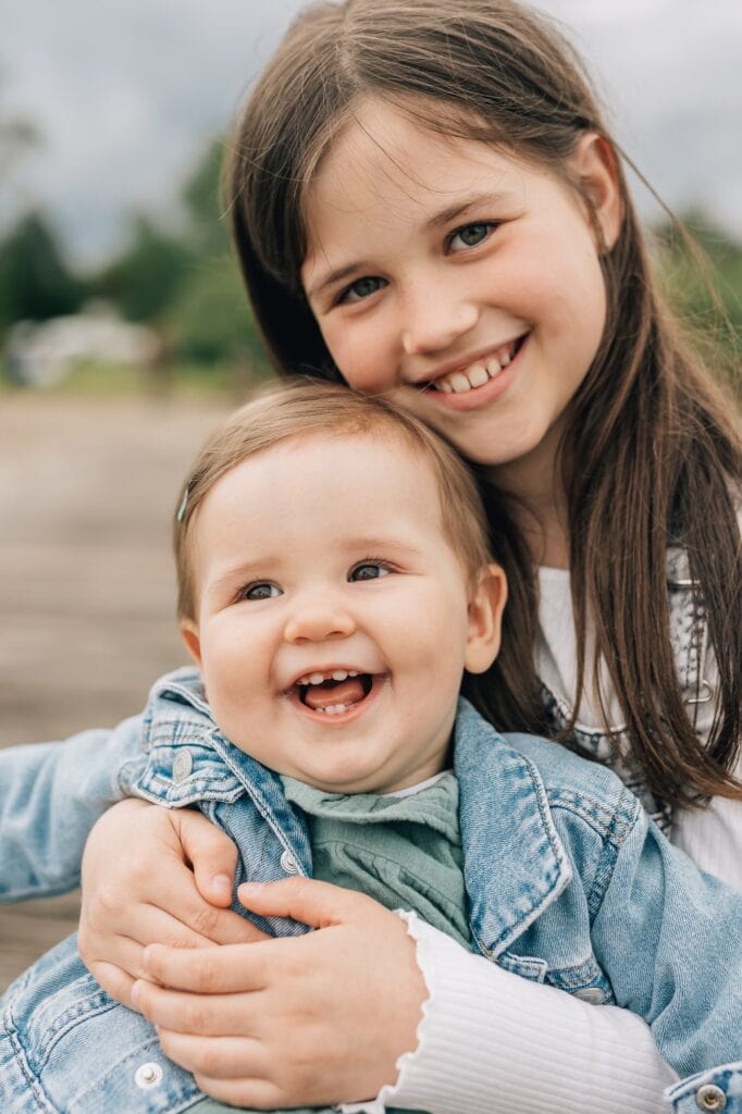 Ein junges Mädchen mit langen Haaren hält ein lächelndes Baby, beide tragen Jeansjacken, Szene im Freien mit unscharfem grünen Hintergrund.