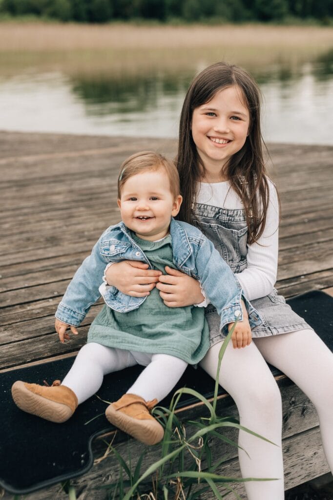 Zwei kleine Kinder sitzen auf einem Holzsteg an einem See. Das ältere Kind, das ein Kleid und ein langärmeliges Hemd trägt, hält ein lächelndes Kleinkind, das eine Jeansjacke und ein grünes Kleid trägt.