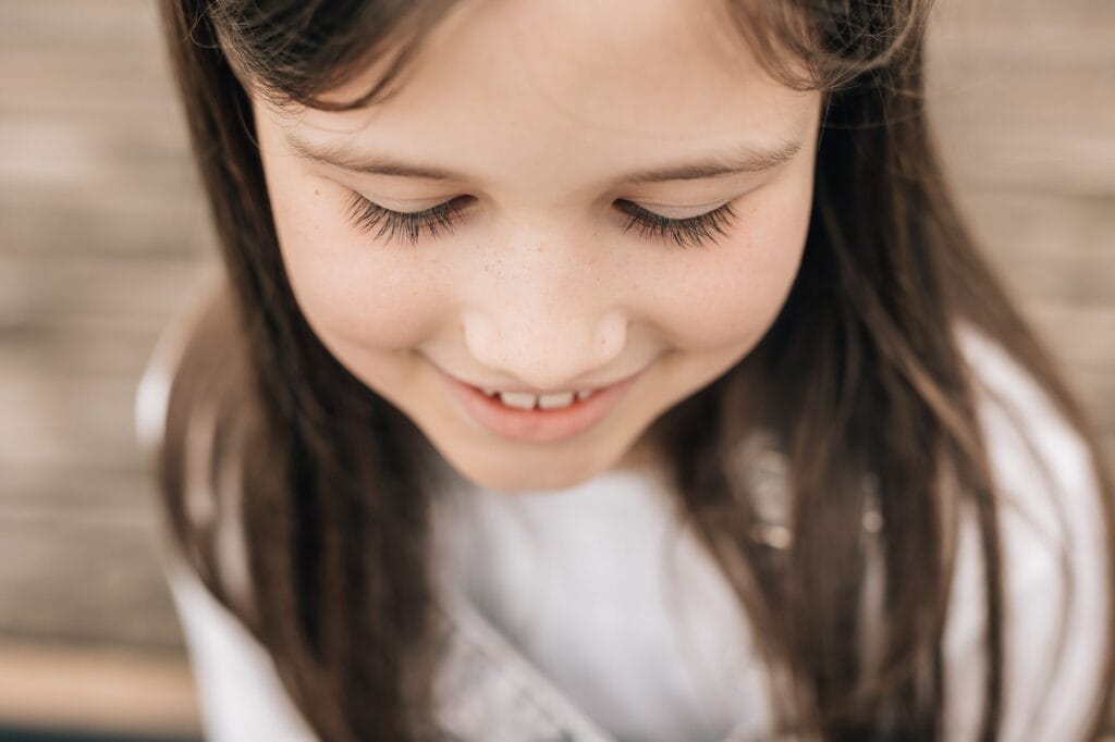 Nahaufnahme eines jungen Mädchens mit braunem Haar, das lächelt und nach unten schaut, sichtbar von den Schultern aufwärts. Sie trägt ein weißes Hemd.